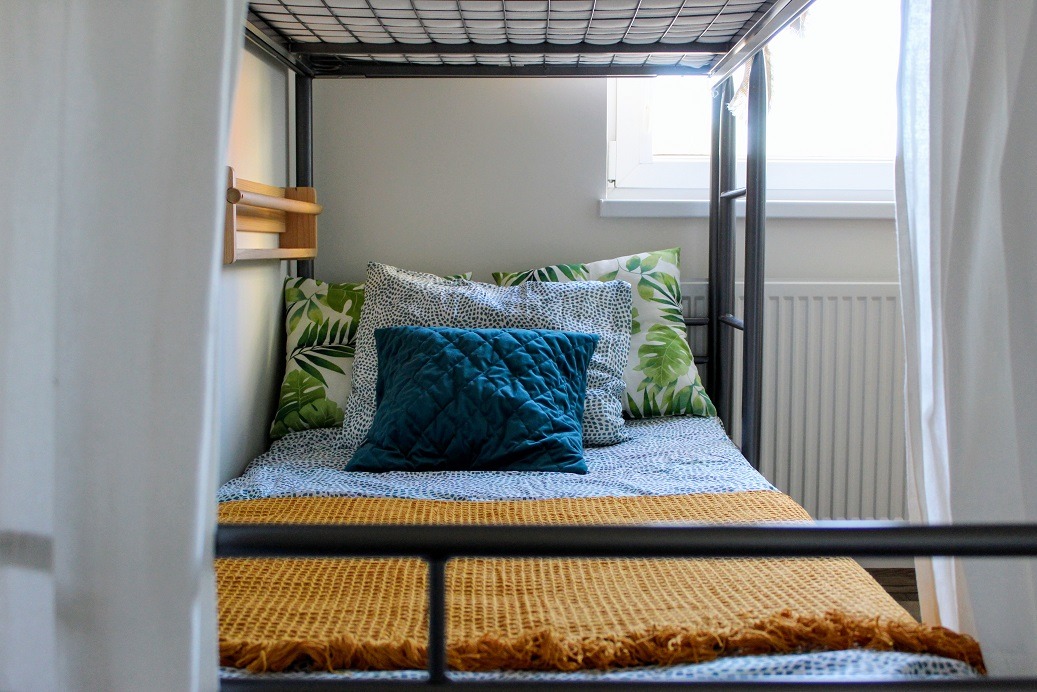 Metalowe łóżko piętrowe z poduszkami w tropikalne liście i turkusową poduszką oraz musztardowym pledem, drewniana półka na ścianie, zasłona i okno w tle.