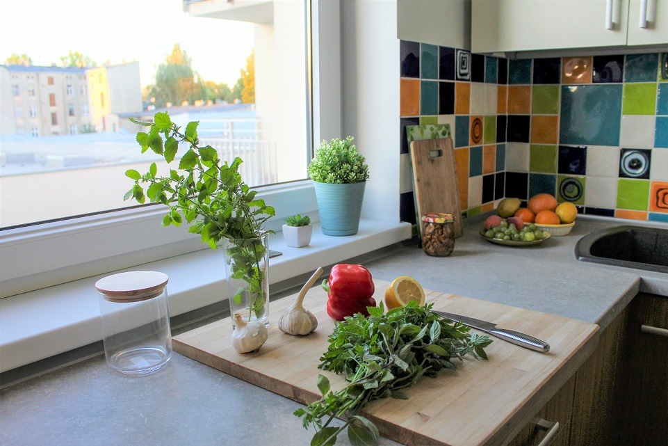 Jasna kuchnia z oknem, zioła w szklance i słoiku na parapecie, deska do krojenia z czosnkiem, papryką, cytryną i ziołami, kolorowe płytki na ścianie.