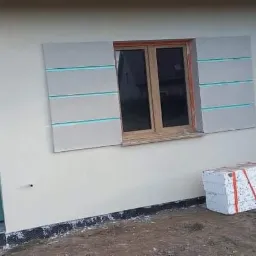 Ściana budynku z częściowo zamontowaną izolacją wokół okna, widoczne bloki styropianowe na ziemi i tymczasowe drzwi wejściowe.