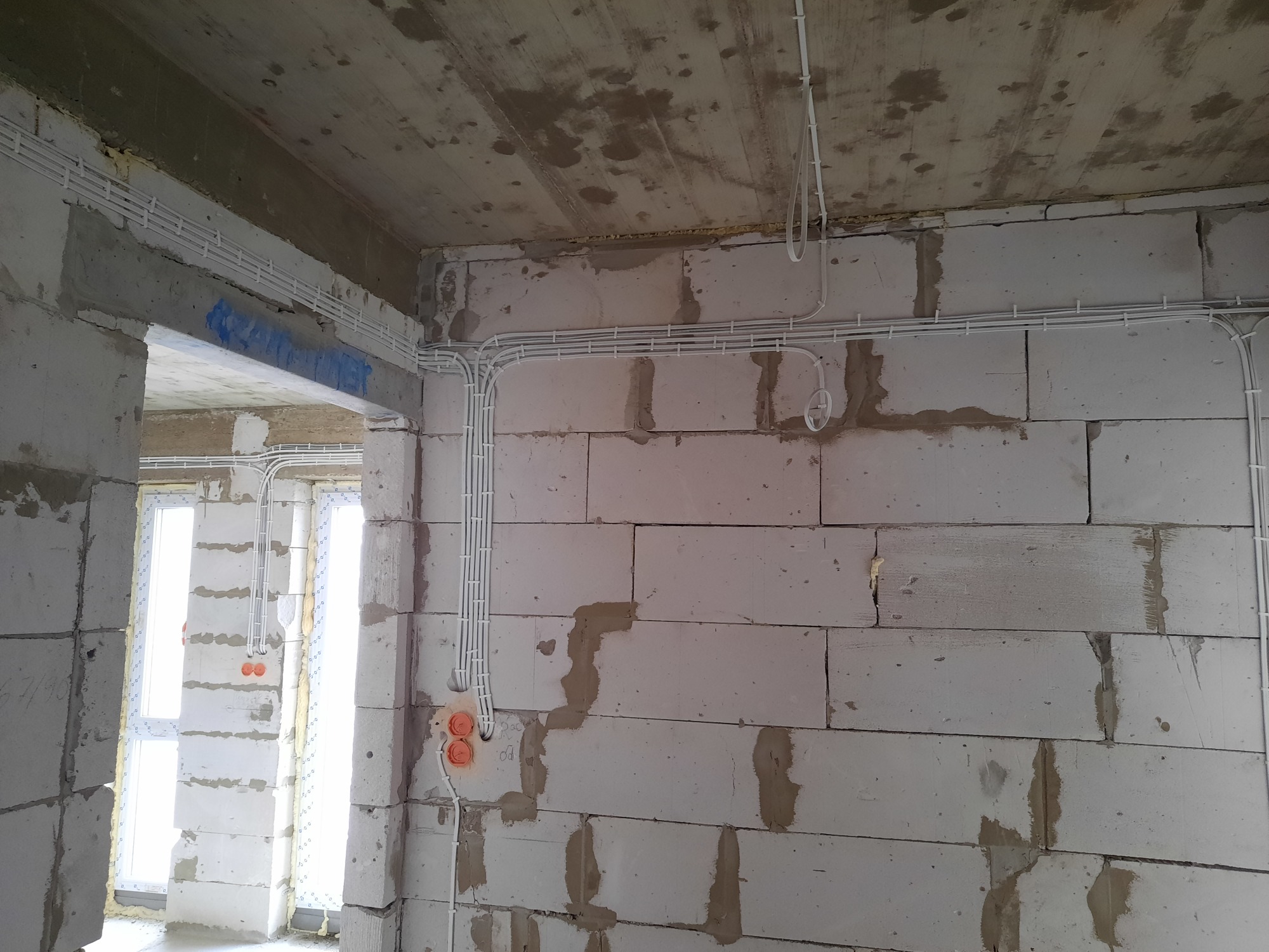 Surowe wnętrze w trakcie instalacji elektrycznej: białe kable prowadzone po ścianach z betonu komórkowego i suficie, widoczne puszki instalacyjne w kolorze pomarańczowym, surowy betonowy strop.