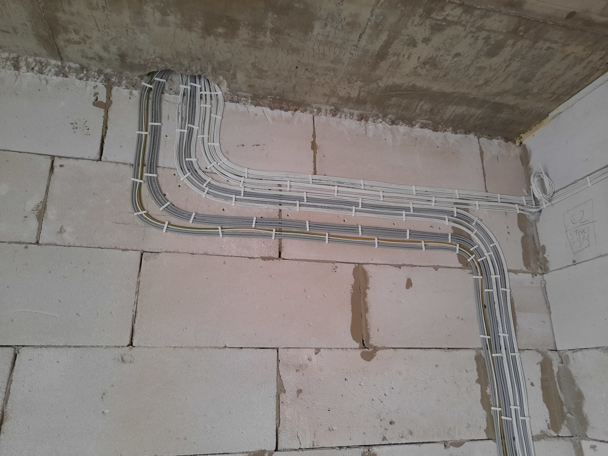 Równolegle ułożone kable elektryczne, przymocowane do ściany z betonu komórkowego za pomocą białych opasek zaciskowych, widoczne pod betonowym sufitem w surowym stanie.
