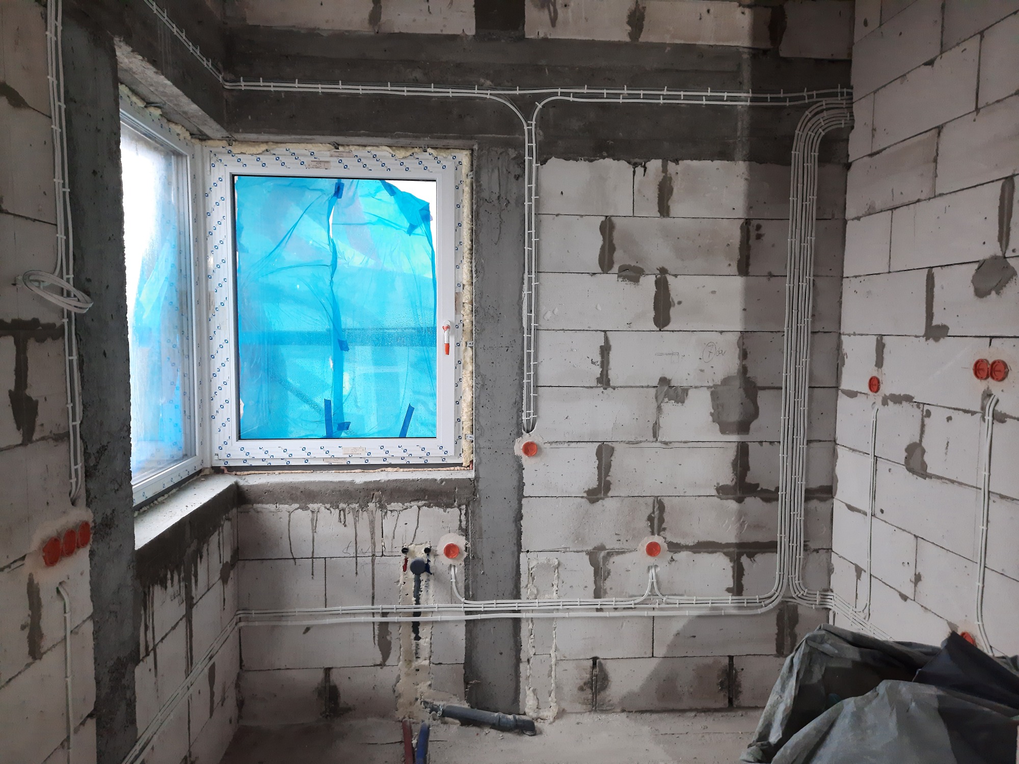 Surowy stan wnętrza z widoczną instalacją elektryczną w białych listwach, okno zabezpieczone folią, ściany z betonu komórkowego.