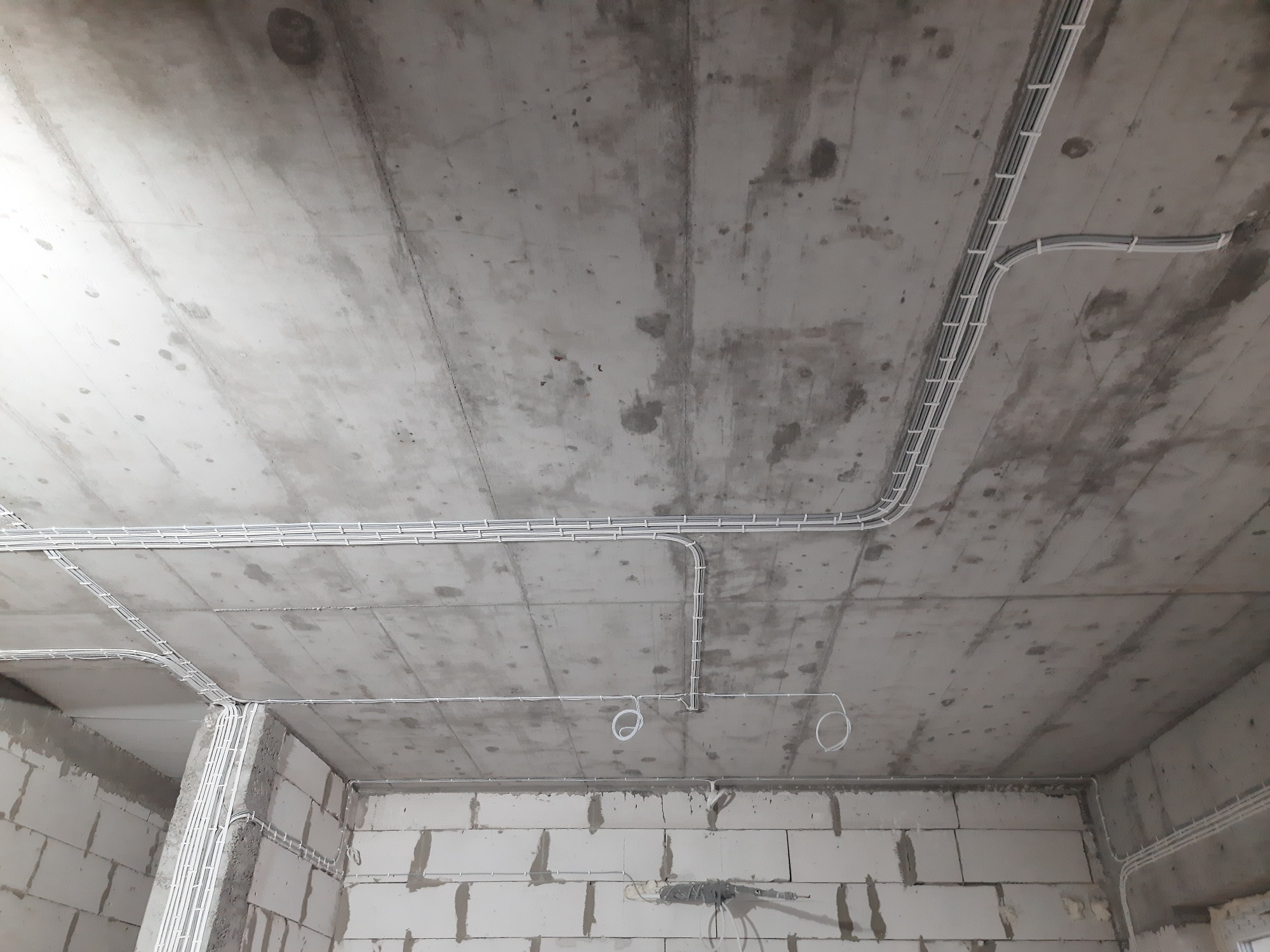 Szare, surowe wnętrze w trakcie prac wykończeniowych z widocznymi, białymi przewodami elektrycznymi ułożonymi w korytkach na suficie i ścianach z betonu komórkowego.