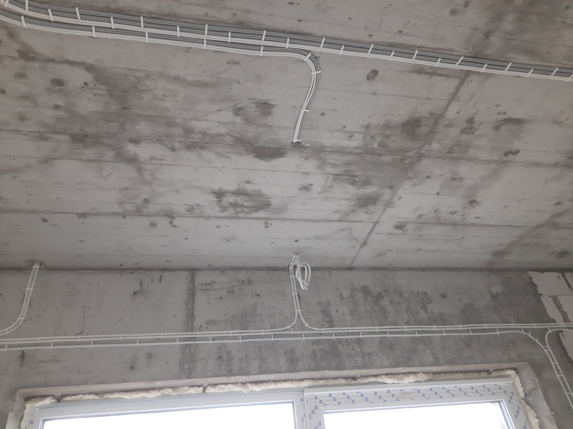 Białe kable elektryczne prowadzone po betonowym suficie w surowym stanie, częściowo w korytach kablowych, widoczne okno w dolnej części kadru.