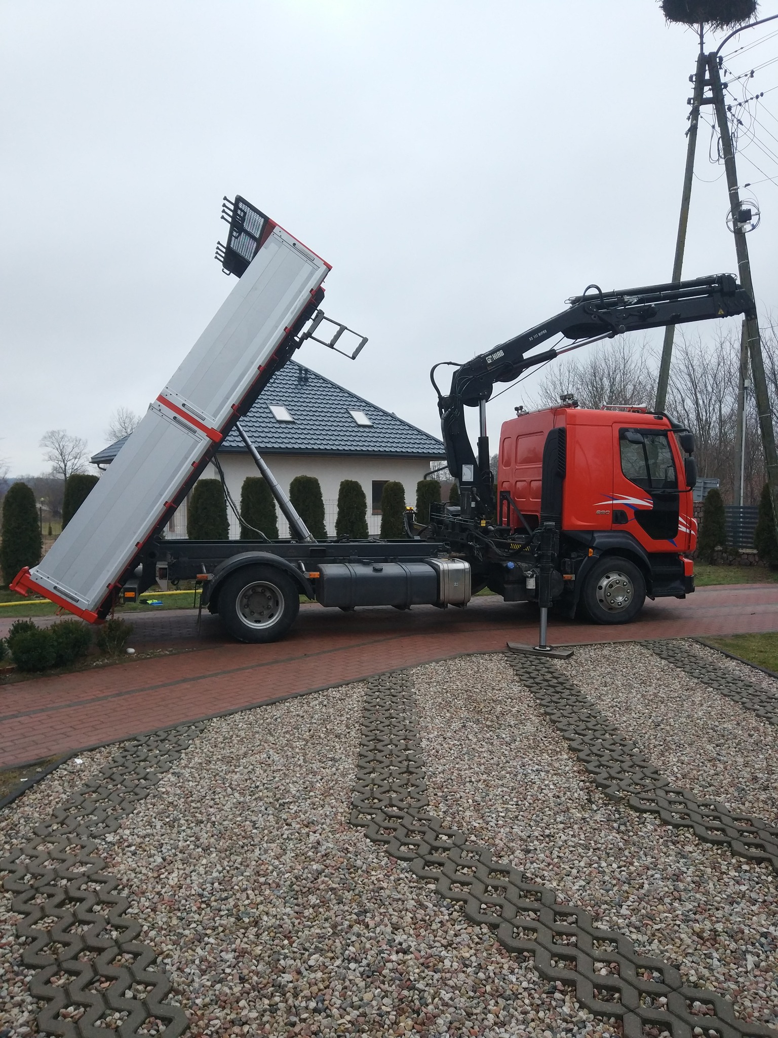 Czerwona ciężarówka z podniesioną skrzynią ładunkową i dźwigiem hydraulicznym na tle domu z szarą dachówką i gniazdem bocianim na słupie.