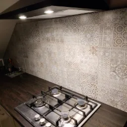 Kuchnia w skosie z płytkami dekoracyjnymi nad blatem i kuchenką gazową, oświetlona lampami okapowymi.