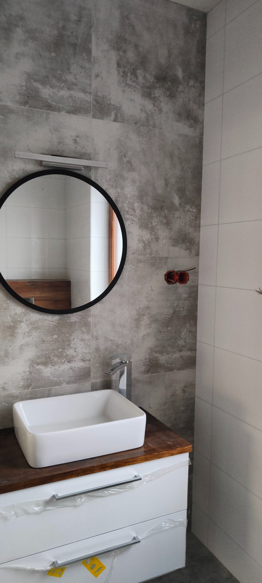 Nowoczesna łazienka w trakcie wykańczania: okrągłe lustro z podświetleniem nad białą, prostokątną umywalką nablatową na drewnianym blacie, szare płytki imitujące beton na ścianie, widoczne kable...