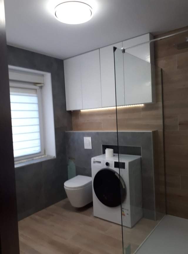 Nowoczesna łazienka po remoncie z drewnopodobną podłogą, szarą ścianą, białą zabudową nad toaletą i pralką, oraz kabiną prysznicową ze szklaną ścianką.