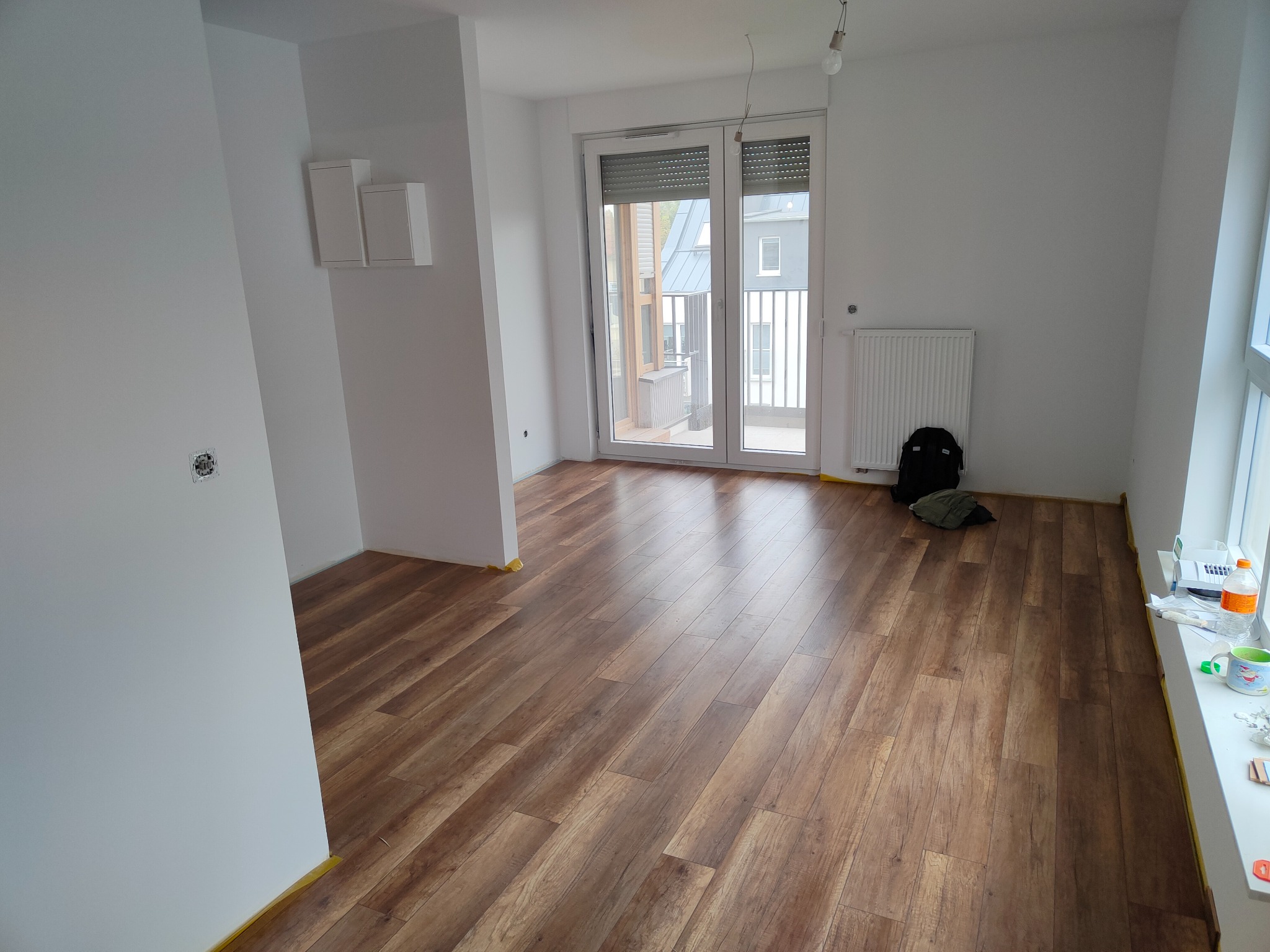 Nowo położona, brązowa podłoga z paneli drewnopodobnych w jasnym, odświeżonym mieszkaniu z widocznymi listwami przypodłogowymi zabezpieczonymi żółtą taśmą malarską.