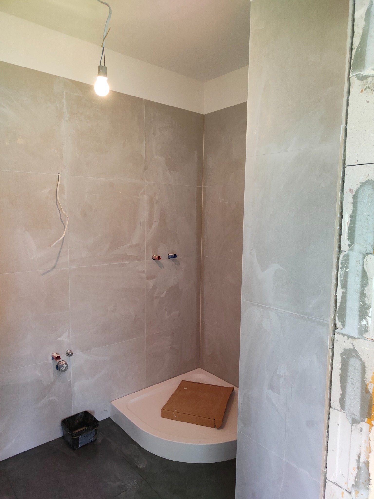 Niedokończona łazienka z szarymi płytkami imitującymi marmur, narożny brodzik z kartonem, widoczne instalacje wodne i wisząca żarówka, ściana z pustaków.