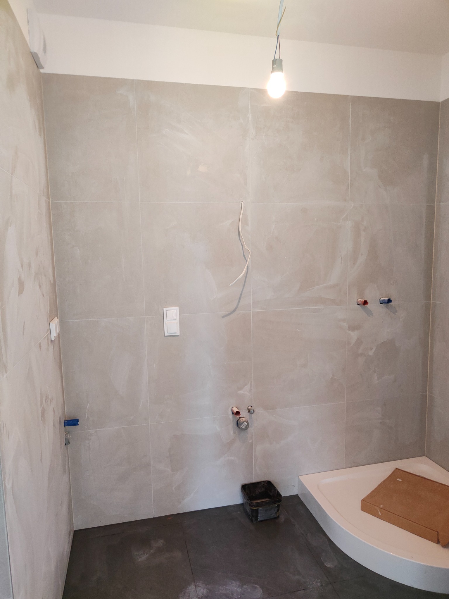 Niedokończona łazienka z beżowymi płytkami ściennymi, ciemną podłogą, brodzikiem z kartonem, wiszącymi kablami i wiadrem na podłodze.