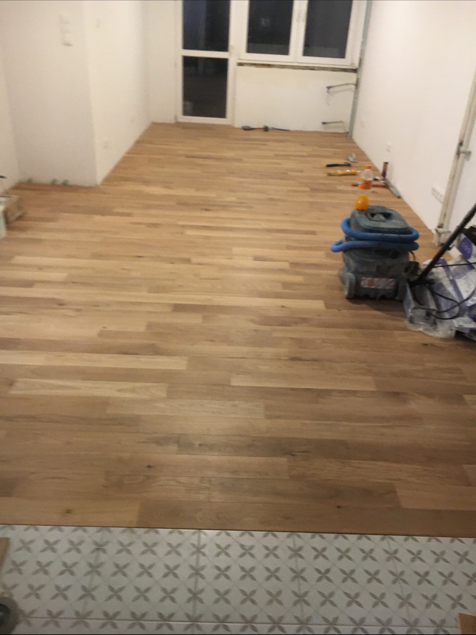 Jasny parkiet ułożony w pokoju, widoczny odkurzacz przemysłowy i narzędzia, dolna krawędź zdjęcia pokazuje fragment podłogi z wzorzystych płytek.