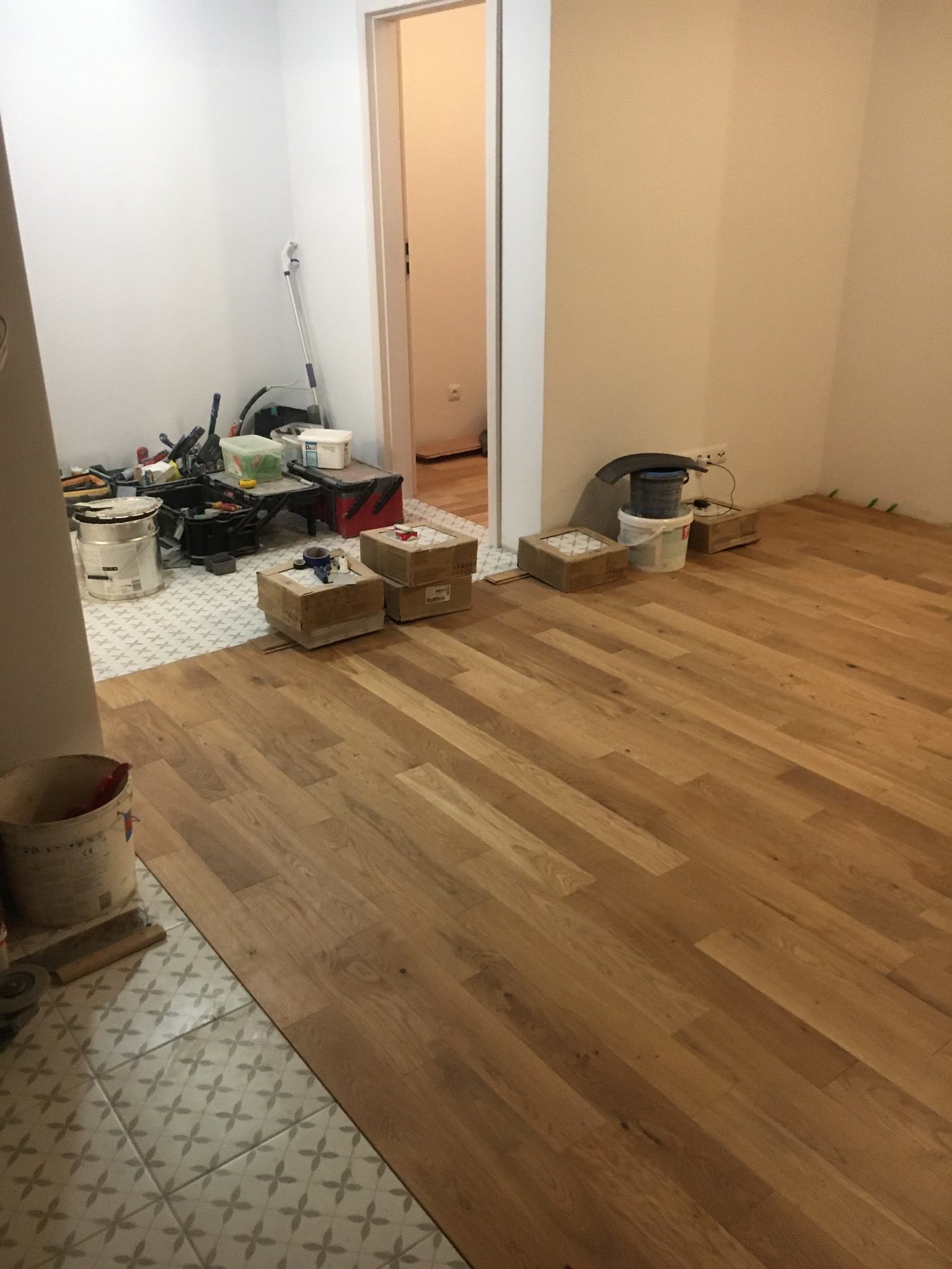 Układanie paneli podłogowych w trakcie remontu: widoczne narzędzia, puszki po farbach i kartony, fragment ułożonych paneli oraz płytek podłogowych w sąsiednim pomieszczeniu.