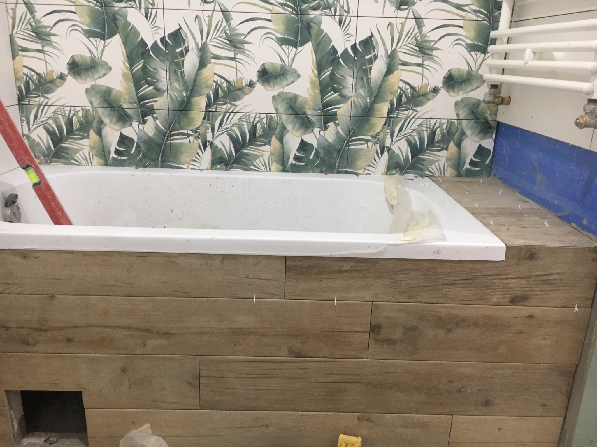 Wanna obudowana płytkami imitującymi drewno, z widocznym poziomującym narzędziem i fragmentem ściany z płytkami w tropikalne liście podczas prac wykończeniowych w łazience.