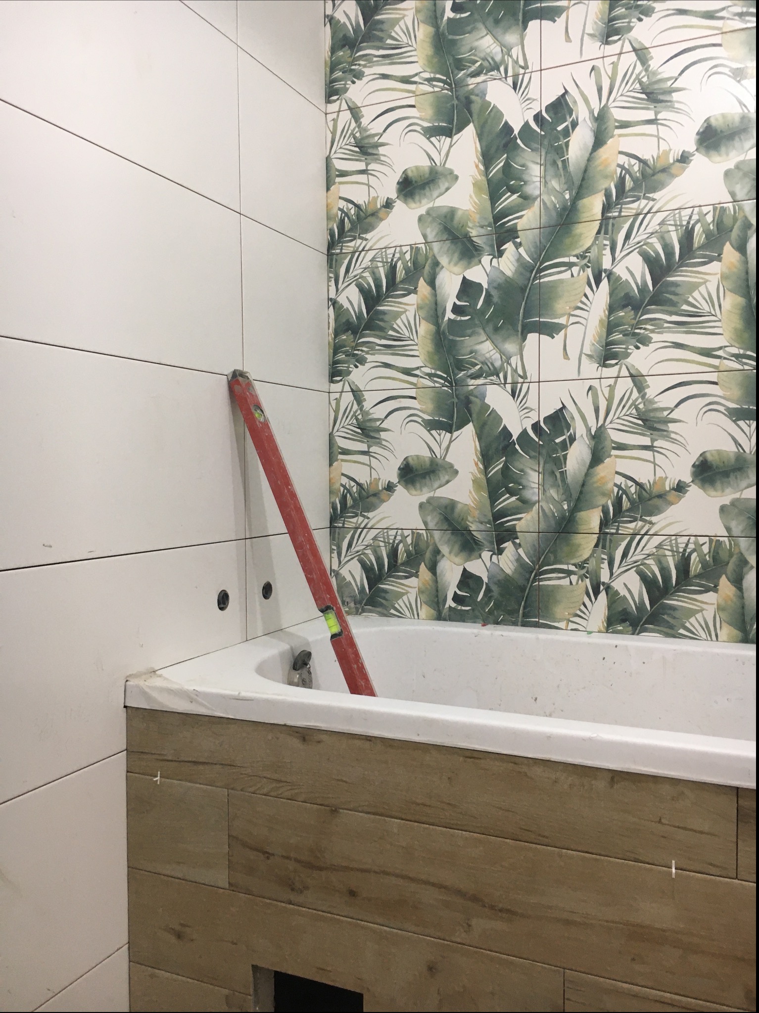 Wnętrze łazienki w trakcie remontu: białe i tropikalne wzory płytek, poziomica przy wannie obłożonej drewnopodobnymi panelami.