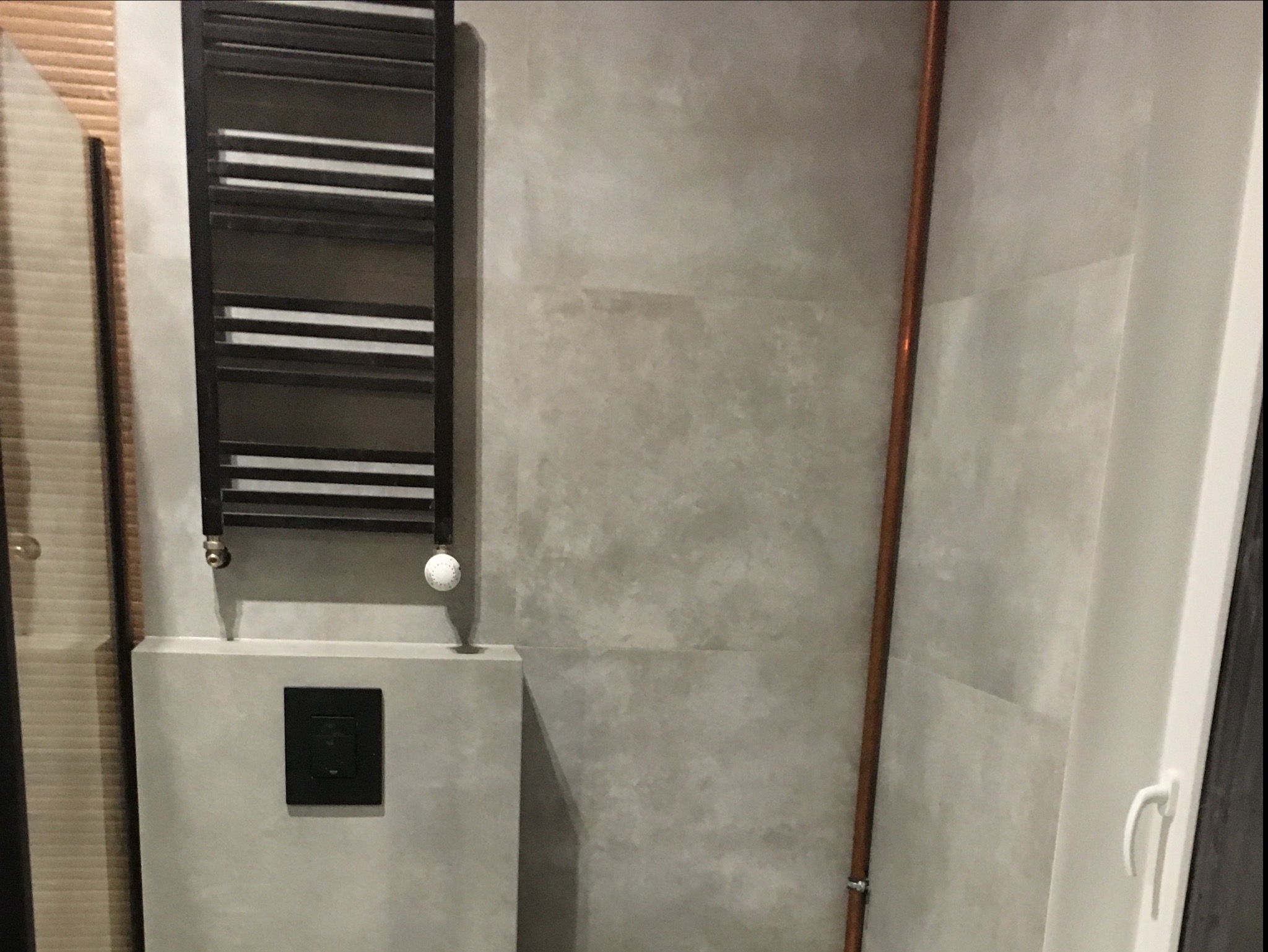 Nowoczesna łazienka z szarą, betonową ścianą, grzejnikiem drabinkowym w kolorze czarnym oraz miedzianą rurą, widoczny fragment okna.