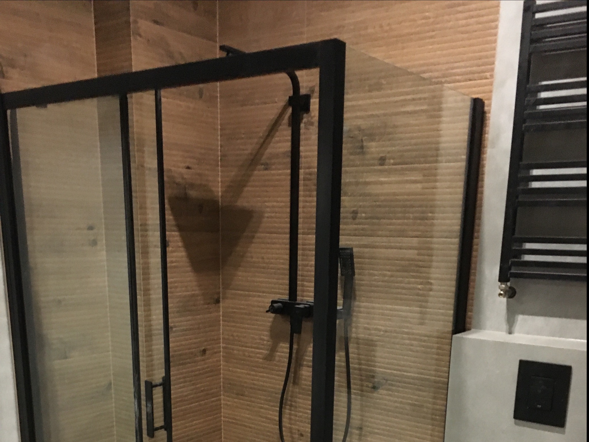 Nowoczesna kabina prysznicowa z czarnymi profilami na tle drewnopodobnych płytek, widoczny czarny grzejnik drabinkowy i minimalistyczna armatura.