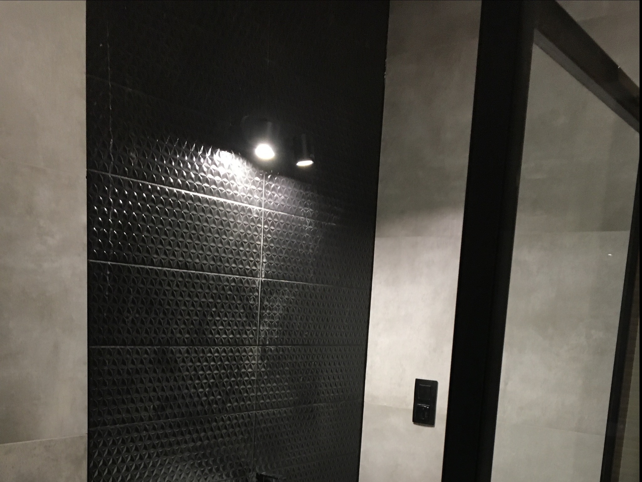 Nowoczesna łazienka z czarnymi, trójwymiarowymi płytkami ściennymi, oświetleniem punktowym i szklaną kabiną prysznicową z czarną ramą.