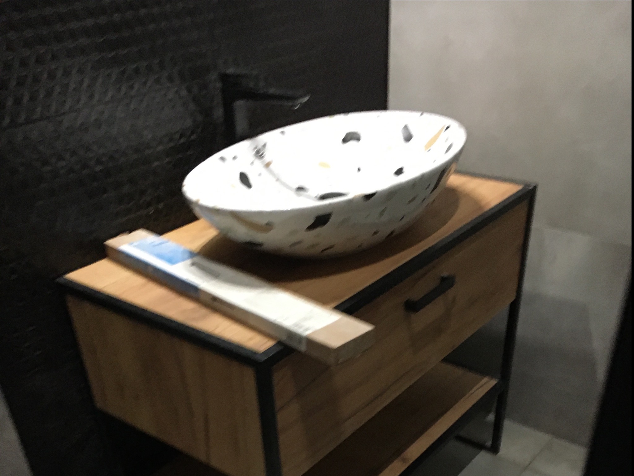 Umywalka nablatowa w stylu lastryko na drewnianej szafce z czarnymi metalowymi nogami, czarna bateria, pudełko z akcesoriami obok, tło z czarnych płytek.