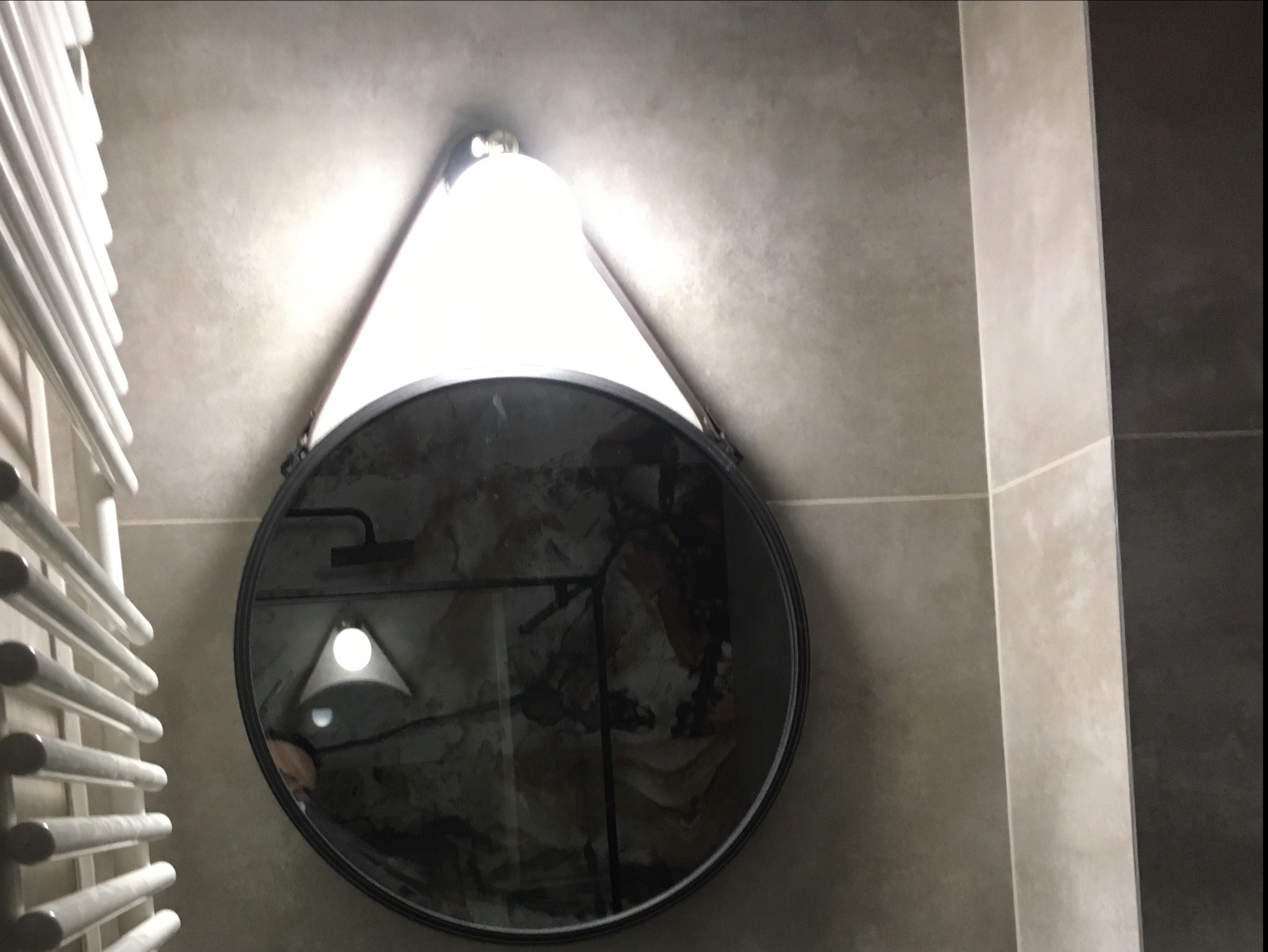 Okrągłe lustro z podświetleniem w łazience, odbijające fragment wnętrza z widoczną armaturą i elementami wykończenia ścian w odcieniach szarości, obok grzejnik drabinkowy.