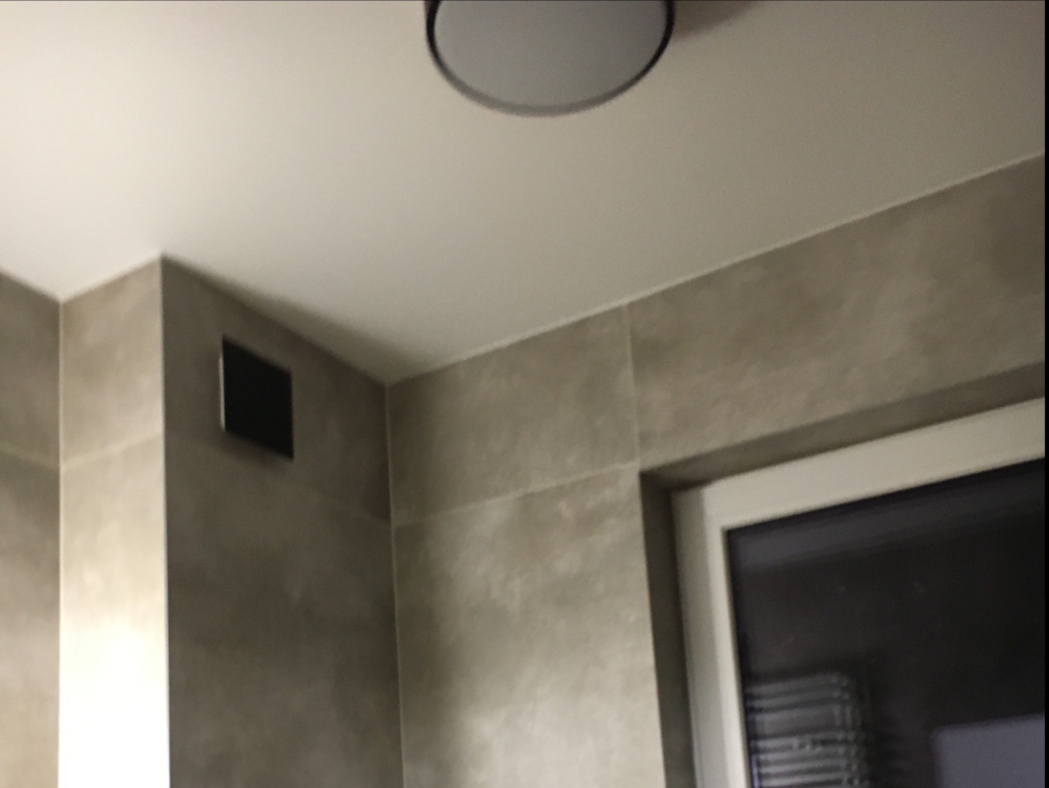 Wykończona łazienka z szarymi płytkami, okrągłą lampą sufitową i kratką wentylacyjną.