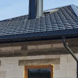 Szare pokrycie dachowe z blachy na nowym domu w trakcie budowy, widoczny komin i okno z pianką montażową.