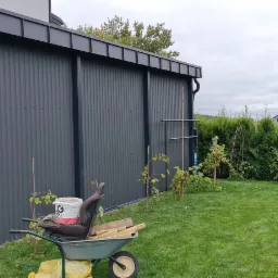 Szara konstrukcja z blachy trapezowej z orynnowaniem i wyposażeniem budowlanym na trawniku, widok na ogród z tujami w tle.