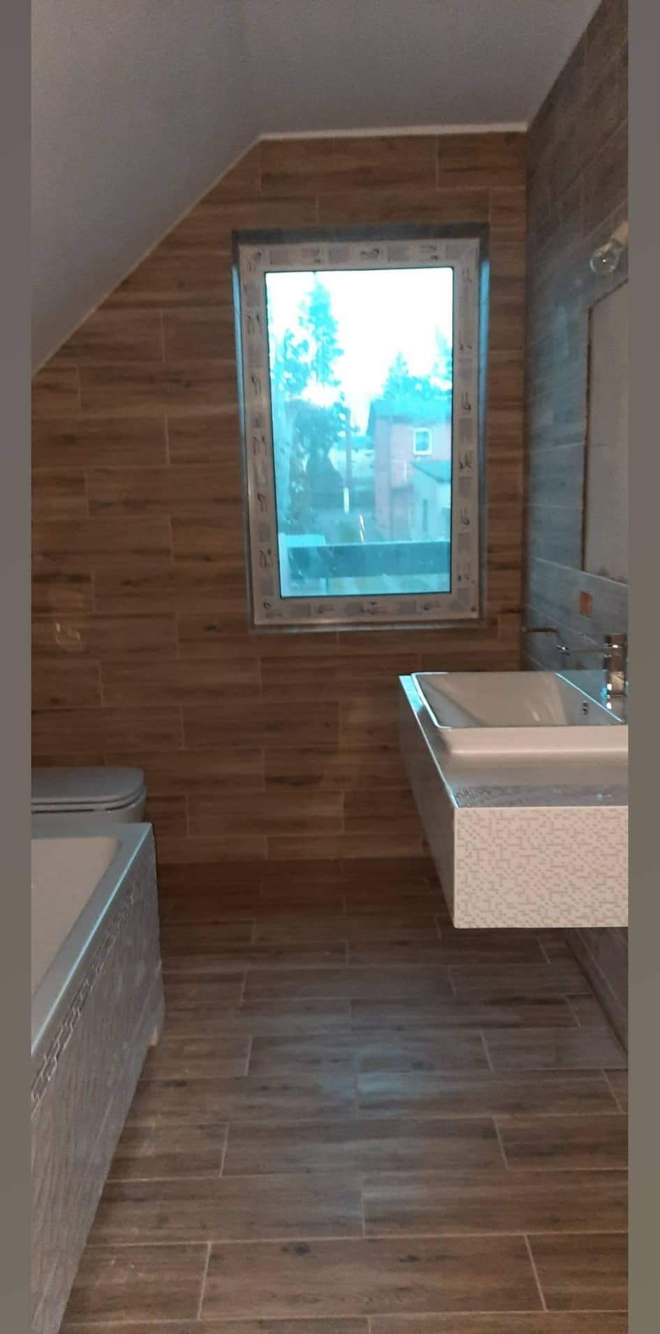 Wykończona łazienka w brązowych odcieniach z drewnopodobnymi płytkami, wisząca umywalka z mozaikową obudową, okno z widokiem na zewnątrz.