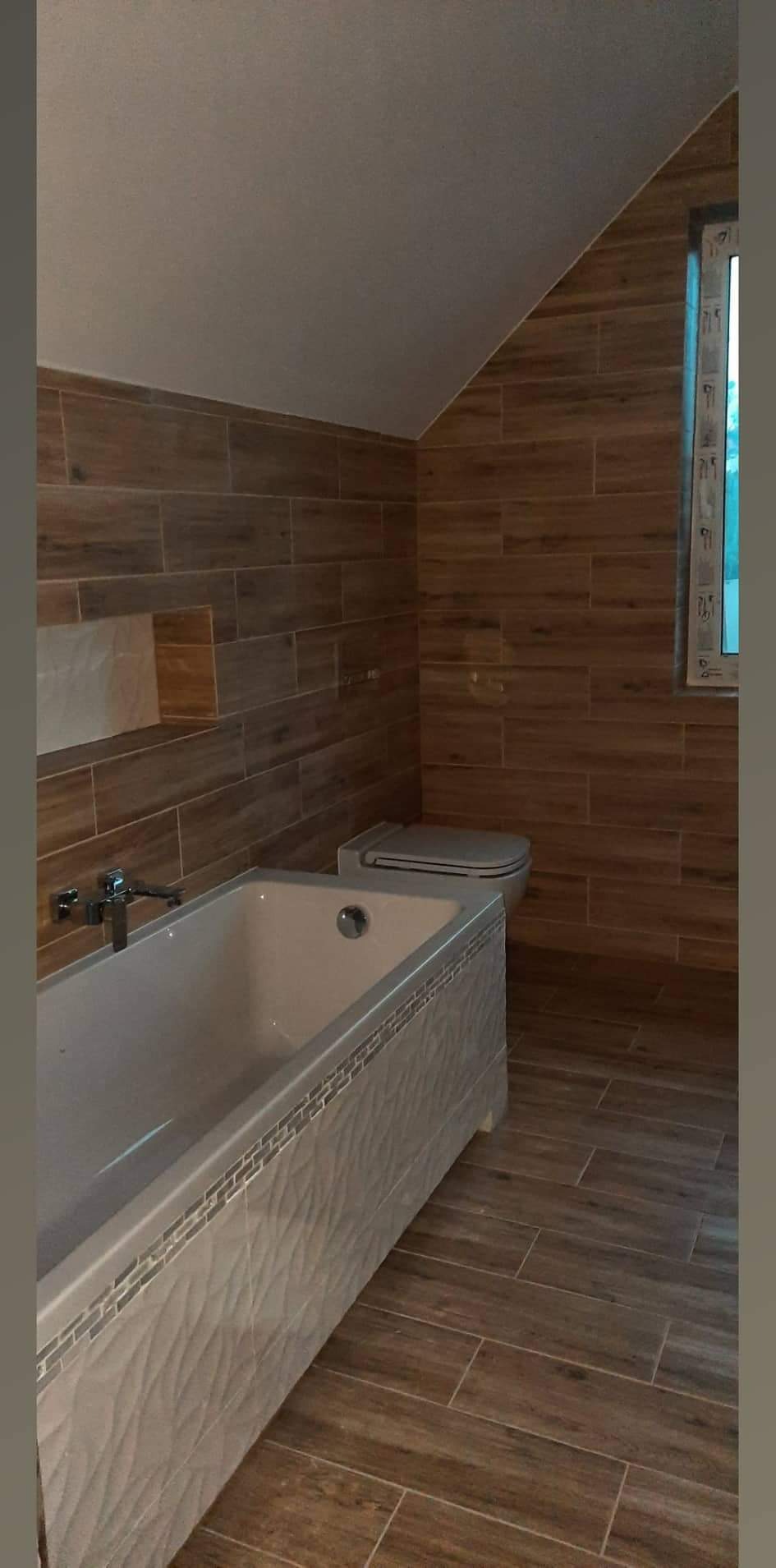 Wykończona łazienka na poddaszu z drewnopodobnymi płytkami na ścianach i podłodze, biała wanna z dekoracyjną obudową i toaleta w rogu przy oknie.