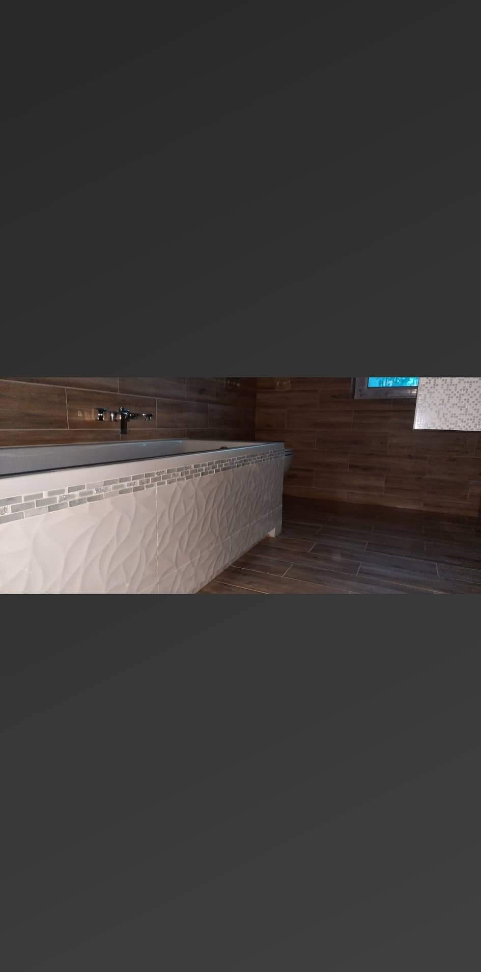 Nowoczesna obudowa wanny z białych płytek o falującej strukturze, wykończona mozaiką, na tle drewnopodobnych płytek ściennych i podłogowych w łazience.