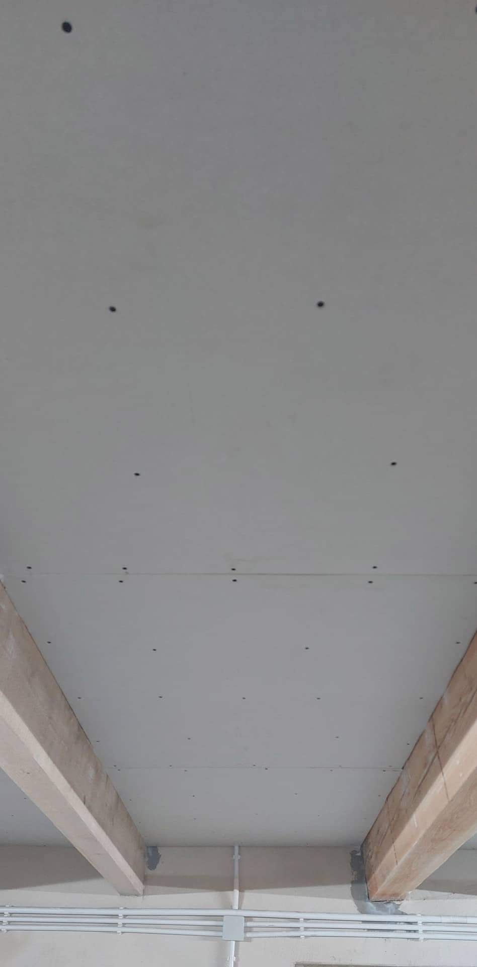 Montaż płyt gipsowo-kartonowych na suficie z widocznymi drewnianymi belkami nośnymi i instalacją elektryczną w tle.