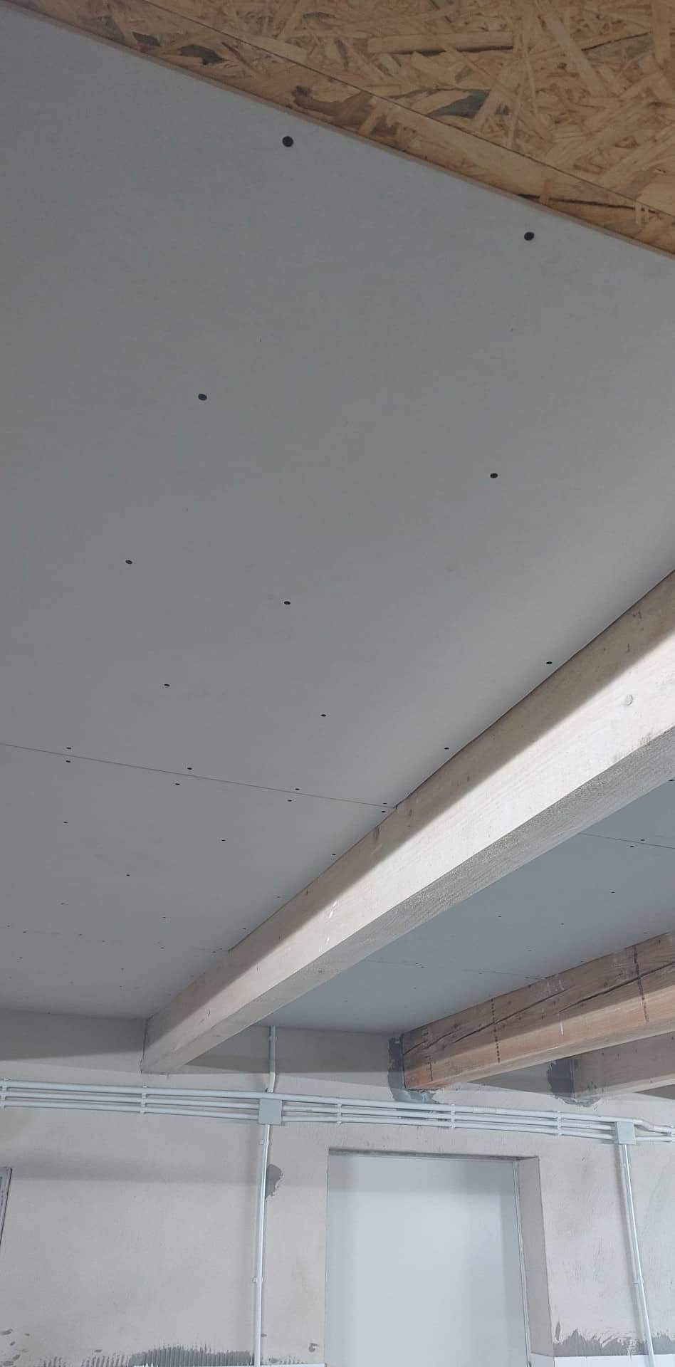 Montaż sufitu podwieszanego z płyt gipsowo-kartonowych, widoczne drewniane belki konstrukcyjne i instalacja elektryczna w peszlach.