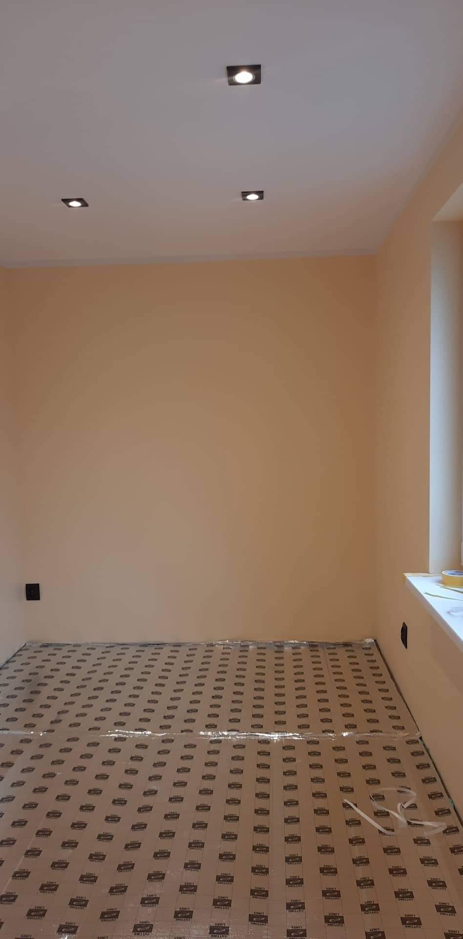 Pokój w trakcie przygotowań do prac wykończeniowych, podłoga zabezpieczona kartonem i folią, ściany pomalowane na ciepły odcień beżu, w suficie zamontowane cztery kwadratowe lampy punktowe.