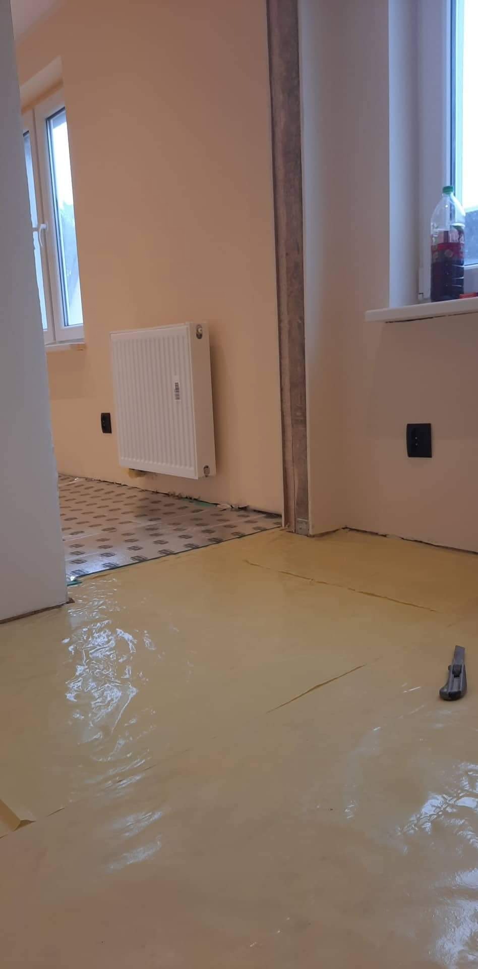 Przygotowanie do remontu: podkład pod panele podłogowe na podłodze zabezpieczonej folią, widoczny grzejnik na ścianie i fragment okna z butelką.