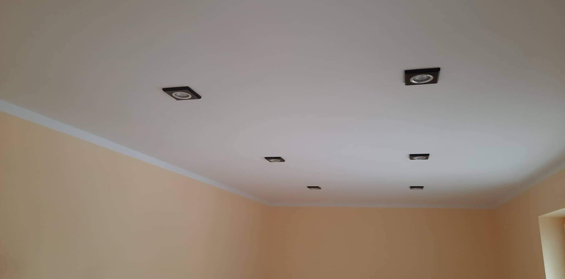 Biały sufit z zamontowanymi kwadratowymi oprawami oświetleniowymi w ciemnych obudowach, widoczne listwy przysufitowe i fragment ściany w odcieniach beżu.