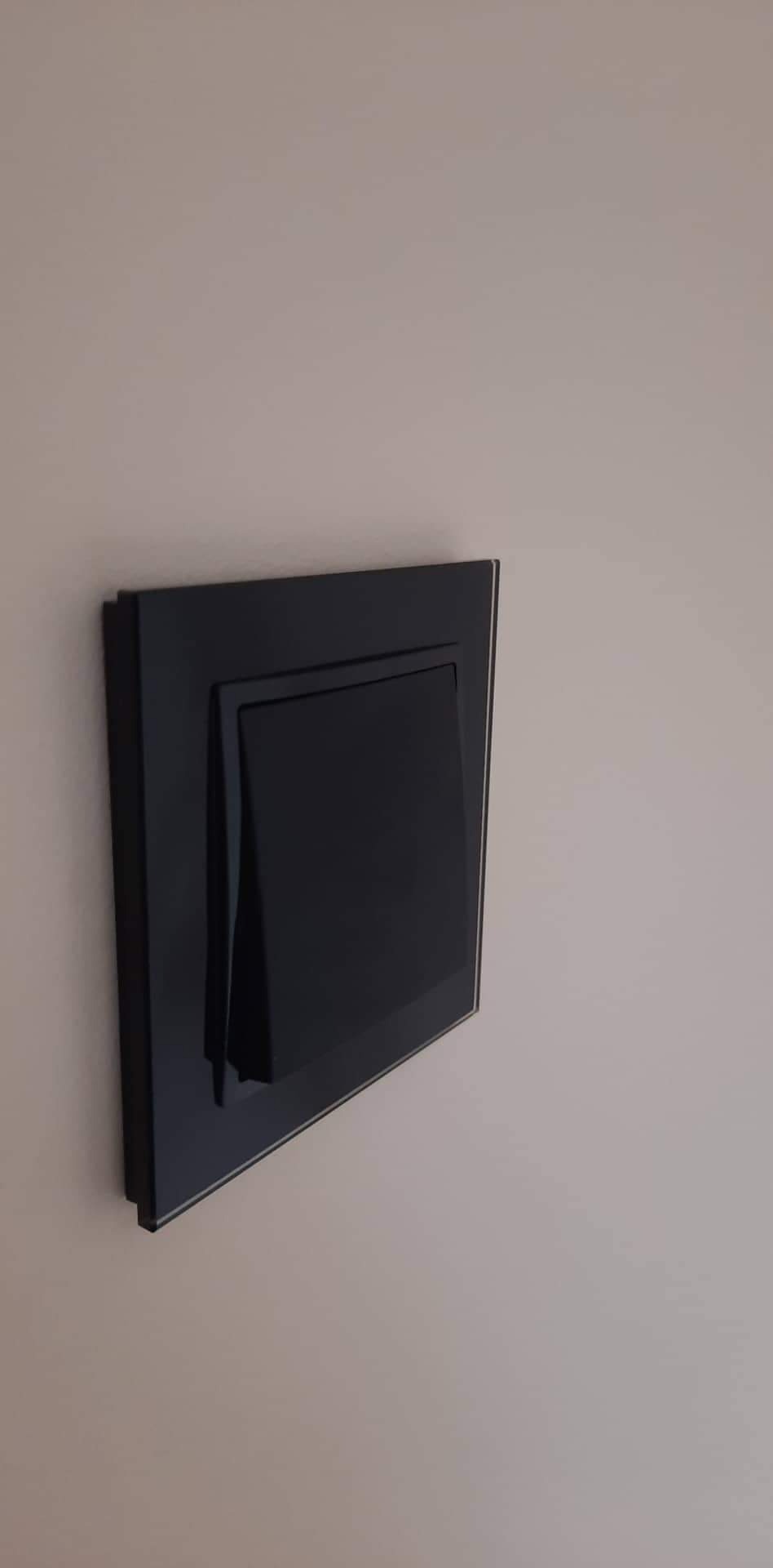 Czarny, kwadratowy włącznik światła z cienką srebrną obwódką, zamontowany na gładkiej, beżowej ścianie, widziany pod kątem.