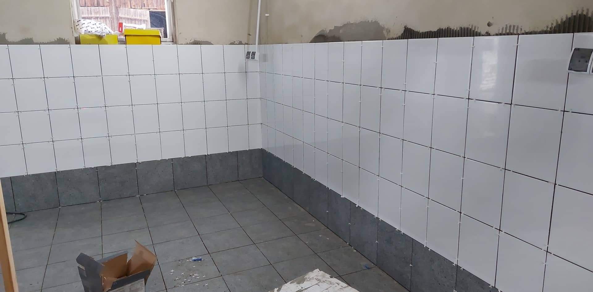 Świeżo ułożone białe płytki ścienne z krzyżykami dystansowymi, kontrastujące z szarymi płytkami podłogowymi i dolnym rzędem szarych płytek na ścianie, widoczne kartonowe pudełko na podłodze.