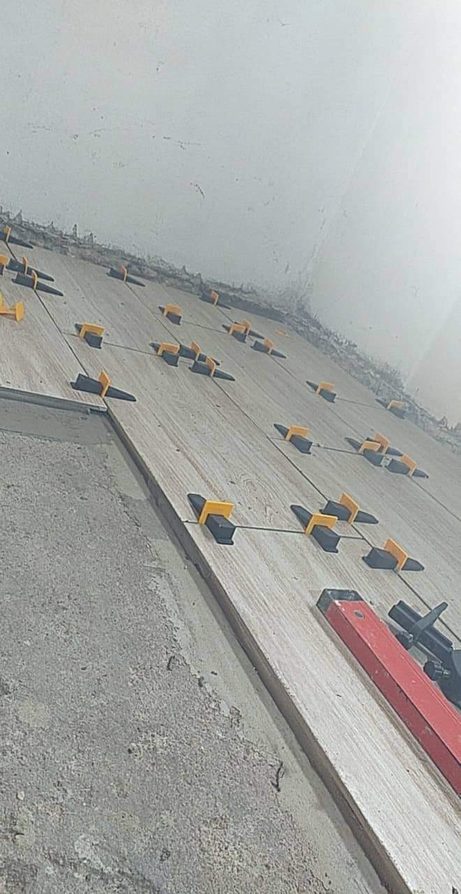 Układanie płytek na ścianie z użyciem klipsów poziomujących i poziomicy, widoczny surowy beton i niedokończona góra ściany.