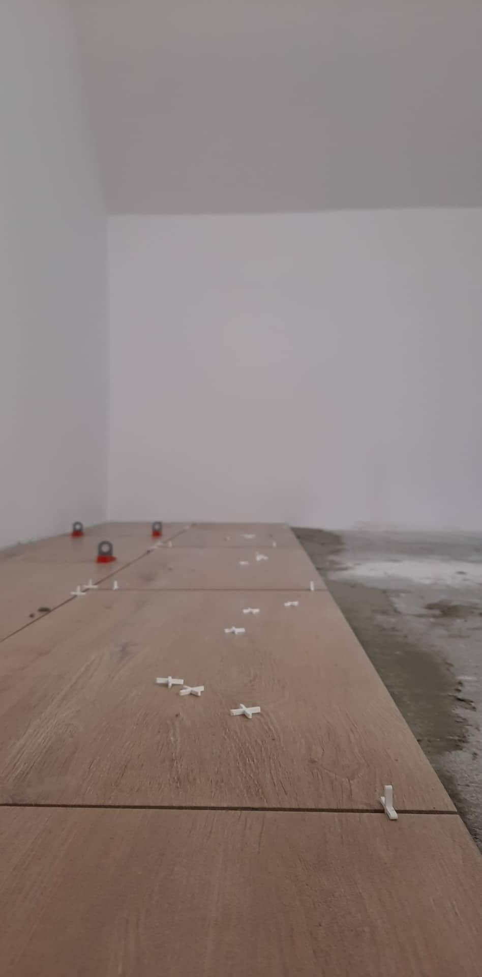 Układanie podłogi z płytek imitujących drewno z użyciem krzyżyków dystansowych i poziomujących klipsów w rogu pomieszczenia.