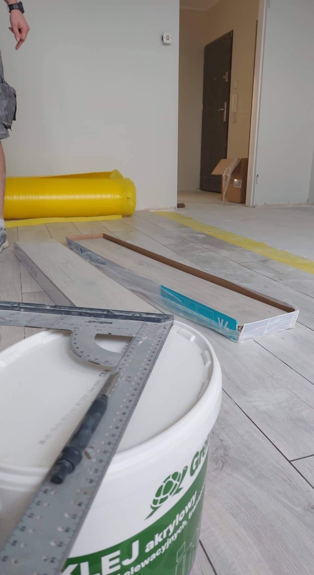 Przygotowanie do montażu paneli podłogowych: wiadro kleju akrylowego, kątownik, paczki paneli w folii ochronnej i folia zabezpieczająca podłoże.
