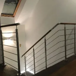 Wewnętrzna klatka schodowa z balustradą ze stali nierdzewnej i drewnianym pochwytem, oświetlona lampami ściennymi i światłem z okna dachowego.