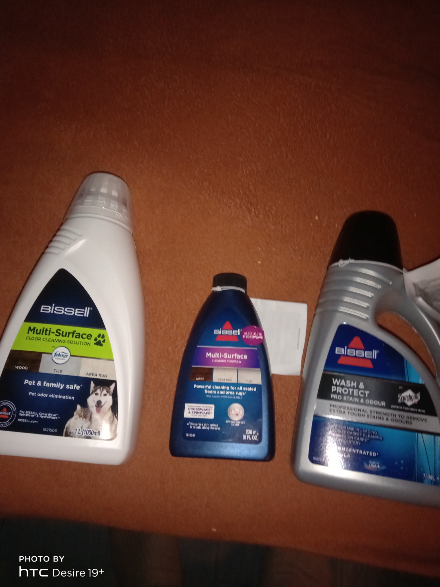 Trzy butelki środków czyszczących Bissell do różnych powierzchni, w tym dywanów, na brązowym tle. Butelki różnią się wielkością i przeznaczeniem, od uniwersalnego płynu do podłóg po preparat...