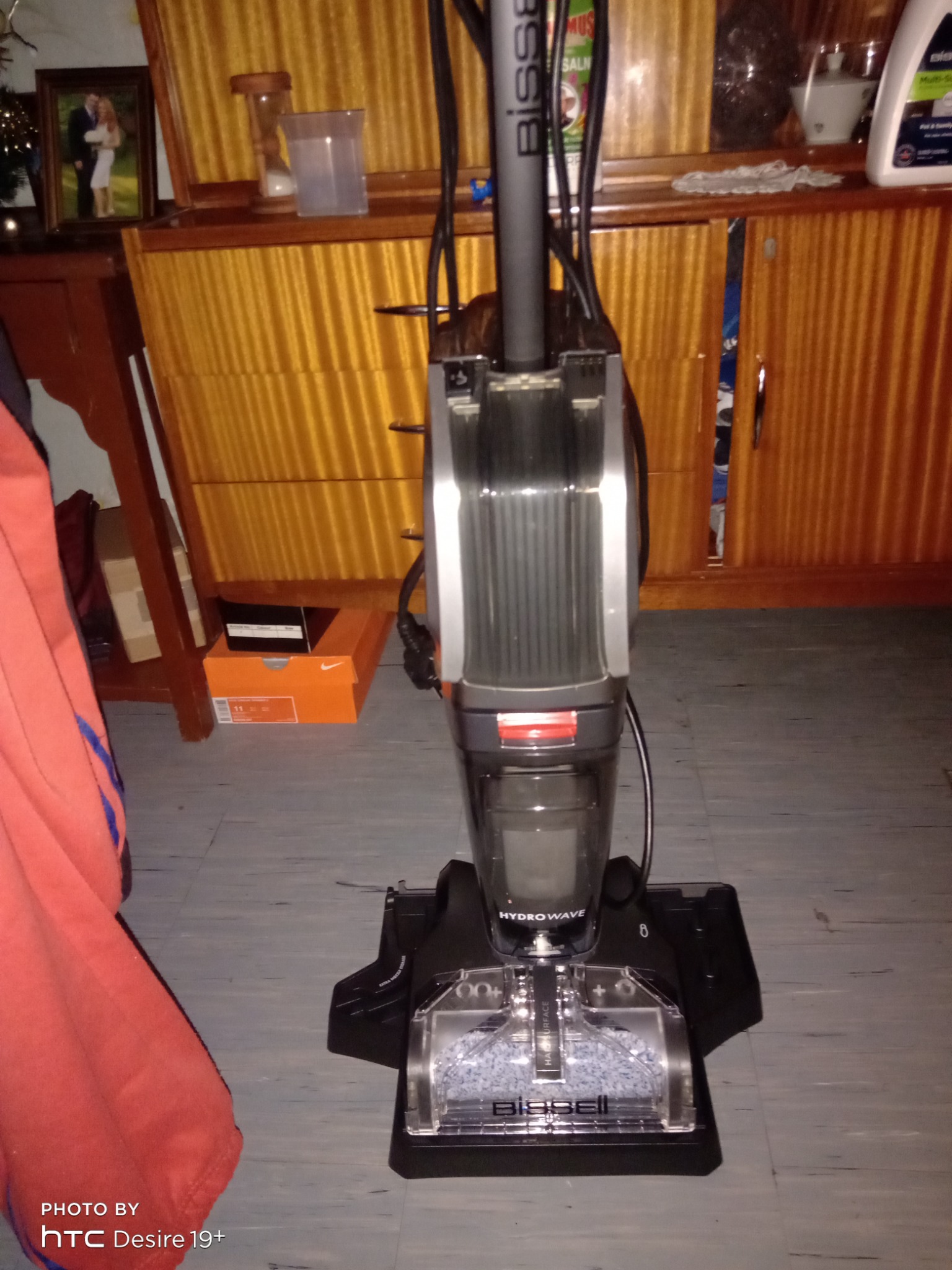 Urządzenie Bissell HydroWave do czyszczenia dywanów i tapicerki na tle drewnianej komody i szarej podłogi.