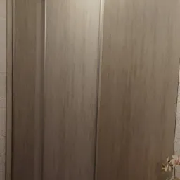 Szafa wnękowa z przesuwnymi drzwiami w odcieniach szarości, z widocznymi metalowymi prowadnicami i kwiatami na stoliku obok.