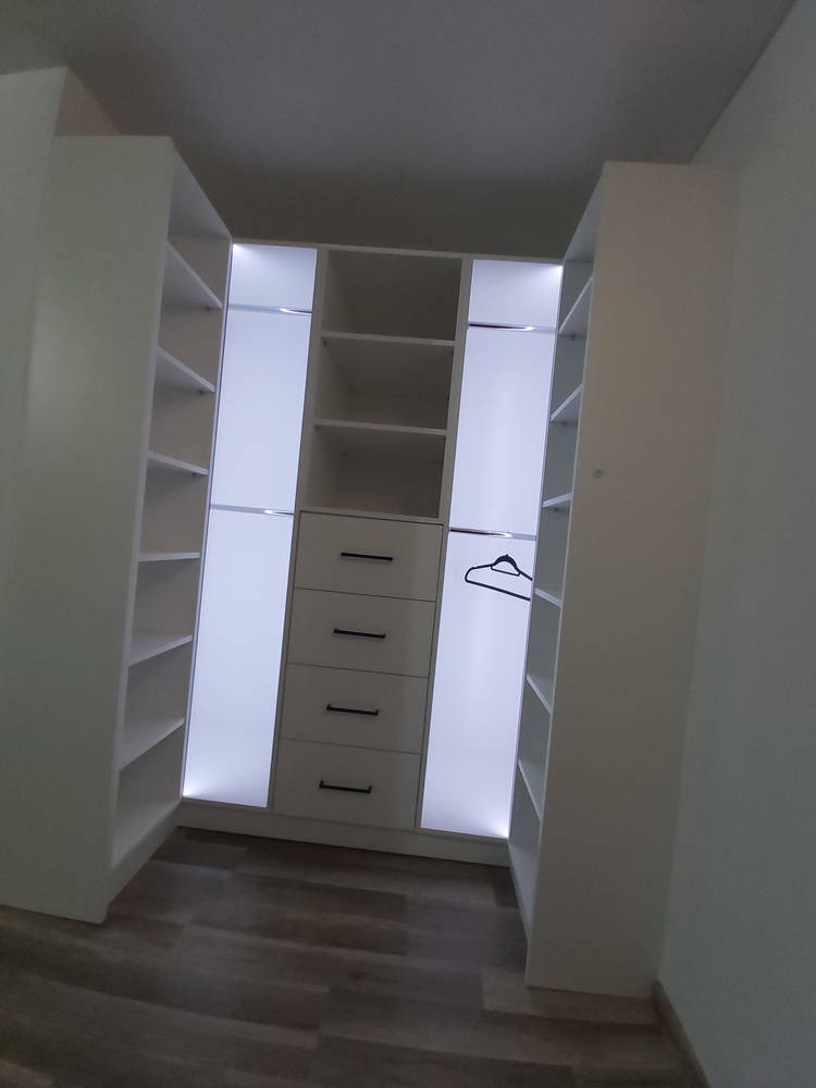 Biała garderoba z szufladami i oświetleniem LED, widok z perspektywy wnętrza, na wieszaku w środkowej części wisi ubranie.