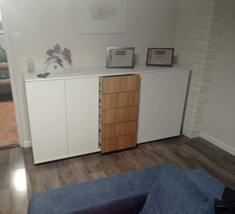 Biała szafka z drewnianymi szufladami, kwiaty i ramki na blacie, ceglana ściana w tle, widok z perspektywy kanapy.