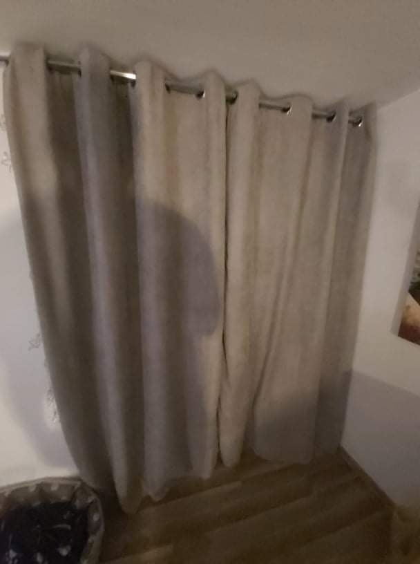 Beżowe, zaciemniające zasłony na metalowym karniszu, z widoczną fakturą materiału i fragmentem parkietu na podłodze.