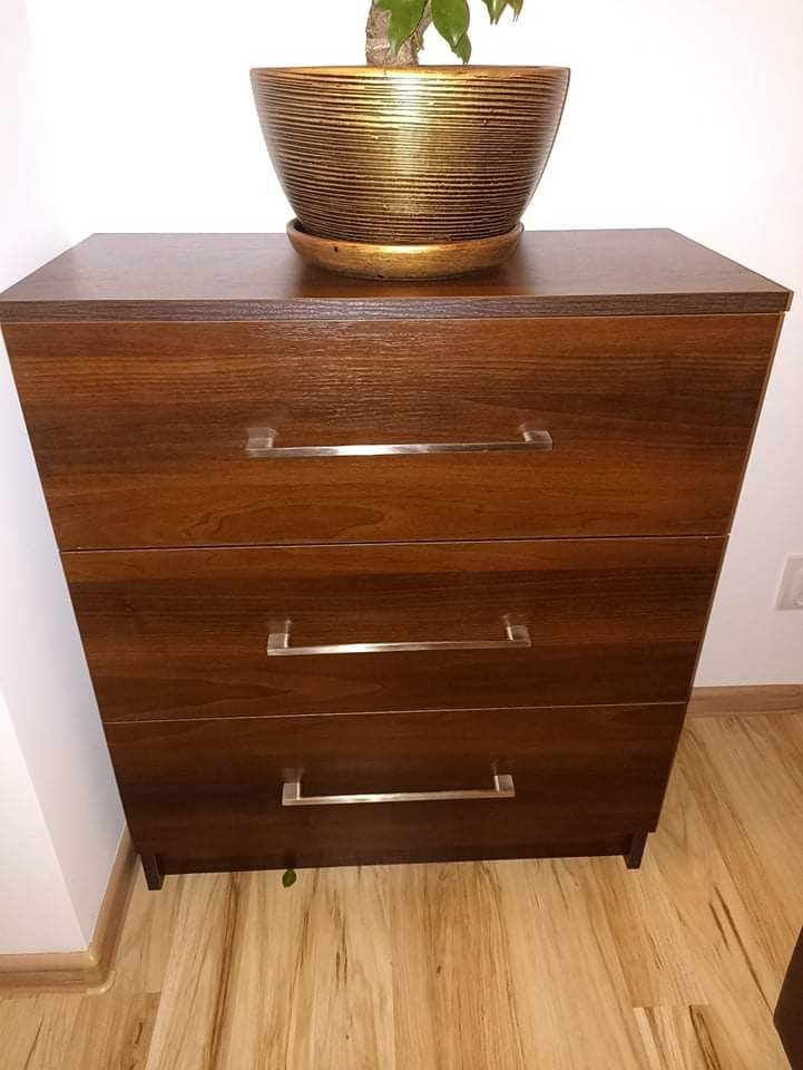 Brązowa komoda z trzema szufladami i srebrnymi uchwytami, na blacie złota doniczka z zieloną rośliną, widoczny fragment podłogi z paneli.