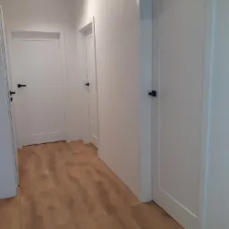 Korytarz z trzema białymi drzwiami z czarnymi klamkami i laminowaną podłogą w odcieniach brązu, oświetlony reflektorami sufitowymi.