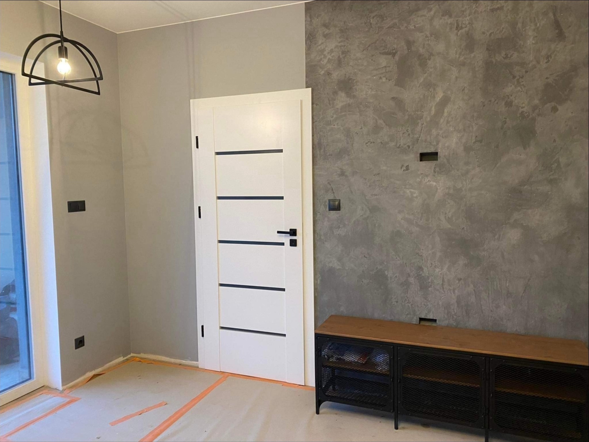 Wnętrze po malowaniu ścian z efektem dekoracyjnym, widoczna biała, nowoczesna stolarka drzwiowa z czarnymi akcentami, zabezpieczona podłoga folią malarską, czarna lampa wisząca i szafka RTV w stylu...